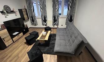 A modern living room with a gray sofa and a wooden table. In the corner stands a black TV on a piece of furniture and curtains with a striking pattern.
