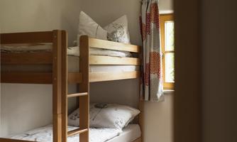 A cozy children's room with a bunk bed and colorful pillows. The room has wooden floors and a colorful carpet pattern.