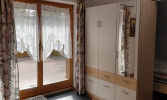 A cozy bedroom with a double bed and a large wardrobe. The windows are adorned with white curtains and let in plenty of daylight.