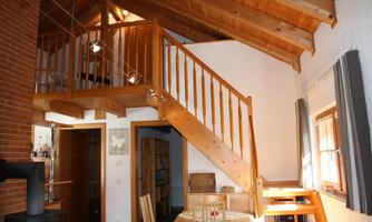 A cozy interior view of a house with a wooden staircase and an open space. The ceiling is made of wood, and there is a dining table in the foreground.