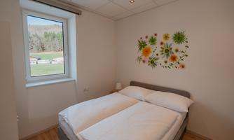 A bright bedroom with a double bed and a colorful flower decoration on the wall. A green landscape is visible through the window.