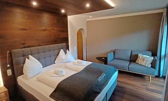 A modern hotel room with a large double bed and a cozy seating area. The walls are designed in warm wooden tones and the lighting creates an inviting atmosphere.
