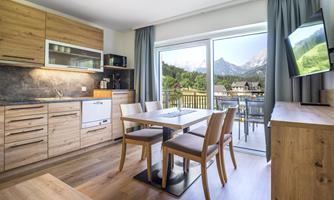 A modern kitchen with wooden cabinets and a dining table. Large windows offer a view of the mountains.