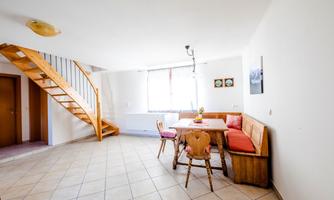 A cozy dining area with a wooden table and benches. A staircase leads to the upper floor, and windows provide plenty of daylight.