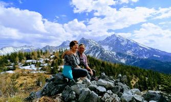 A couple is sitting on a rock in the mountains. In the background, there are snow-capped peaks and a blue sky with some clouds.