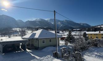 A snow-covered landscape with mountains in the background. In the foreground, there is a house and a bright blue sky can be seen.