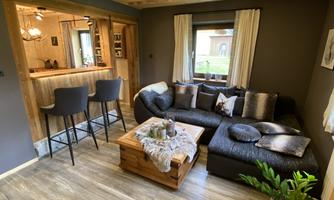 A cozy living room with a dark L-shaped couch and a wooden table. In the background, there is an open bar with high stools and a large window.