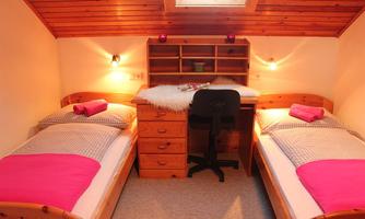 A cozy bedroom with two single beds and a desk. The walls are made of wood, and there is soft lighting as well as decorative pillows in pink.