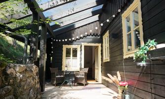 A cozy wooden house with a covered terrace. In front of the house are two chairs and a table, surrounded by plants and decorations.