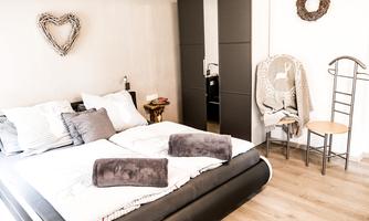 A cozy bedroom with a large bed and simple, modern furniture. The walls are in neutral tones and there are decorative elements made of wood.