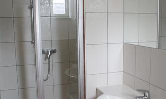 A modern bathroom with a clear shower cubicle and a sink. The walls are tiled in light colors, and there is a large mirror.