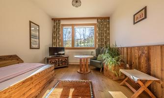 A cozy room with wooden decor and large windows. There is a bed, a television, and a small table with a chair.