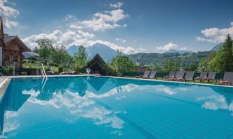 A sparkling pool with clear water and a view of the mountains. Surrounded by loungers and green grass under a blue sky.