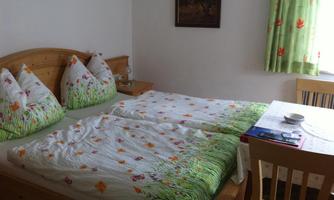 A cozy bedroom with a double bed and colorful floral bedding. The room is bright and friendly with a small table and a window.