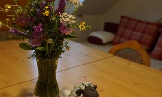 A beautiful table with a vase full of colorful flowers and a cute sheep decoration. In the background, some cozy seating options can be seen.