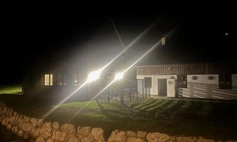 A house at night with bright lights. The garden is visible, surrounded by a stone wall.