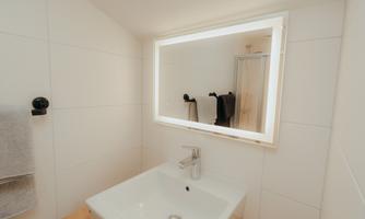 A modern bathroom with a sink and a lit mirror. In the background, towels and a shower cabin can be seen.