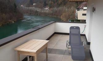A bright terrace with a table and a lounge chair. In the background, a green river flows between wooded hills.