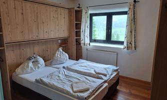A simple bedroom with two beds and wooden shelves. The room has a large window with a view of the greenery.