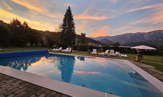 A swimming pool with clear water reflects the colorful sunset. In the background, trees and mountains can be seen.