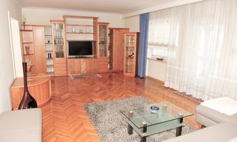 A modern living room with wooden shelves and a television. The room has large windows, bright furniture, and a fluffy rug.