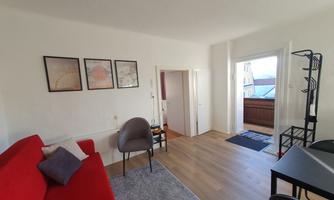 A modern living room with a red sofa and wooden floor. There is space for a table and chairs, as well as access to a balcony.