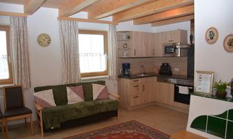A cozy kitchen with wooden shelves and modern appliances. In the foreground, there is a sofa with decorative cushions and a rug on the floor.
