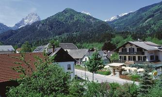 A picturesque village surrounded by high mountains and lush greenery. In the foreground, cozy houses and an inviting café can be seen.