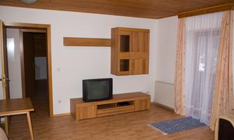 A simple living room with a wooden shelf and a television. Bright walls and a wooden floor create a warm atmosphere.