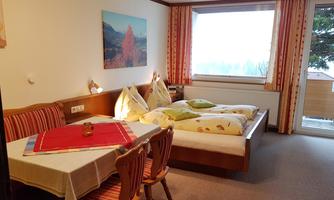 A cozy room with a double bed and a table. Large windows provide a view outside and the walls are decorated with colors and pictures.