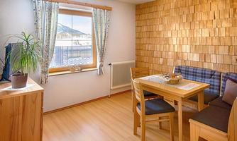 A cozy living space with wooden walls and a large window. There is a table with chairs and a small plant description.