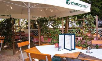 A cozy outdoor restaurant with wooden chairs and a large sunshade. On the table lies a menu, surrounded by green plants.