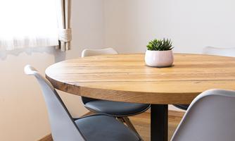 A round wooden table with a small plant pot in the center. Around it are four modern chairs in soft colors.