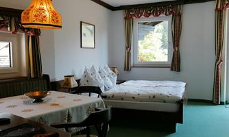 A simple bedroom with a bed and a table. The walls are decorated with patterned curtains and a rustic lamp.