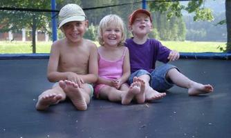 Three happy children are sitting barefoot on a trampoline. In the background, trees and a green meadow can be seen.