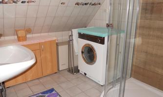 A bathroom with a shower, a sink, and a washing machine. The walls are tiled and there is a colorful rug on the floor.