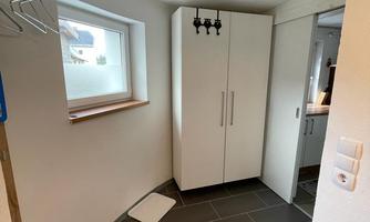 A modern hallway with a white wardrobe and a small window opening. The floor is designed with dark tiles, and there is a simple coat rack on the wall.