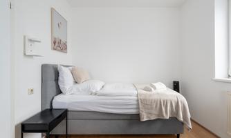 A simple bedroom with a gray bed and a small nightstand. The walls are light and subtly decorated.
