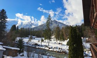 A winter landscape with snow-covered trees and a clear blue sky. In the background, there are tall mountains and a flowing stream.