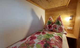 A cozy bed in a wooden room with colorful floral bedding. The walls are light, and a lamp provides soft light.