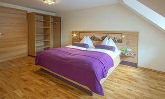 A modern bedroom with a large double bed and purple bedding. The room has wooden flooring and a cozy atmosphere.