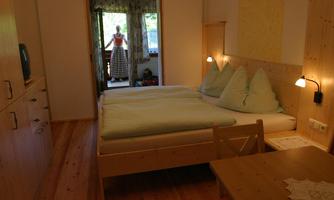 A cozy bedroom with a double bed, light wooden paneling, and a table. In the background, you can see a person standing on a balcony.