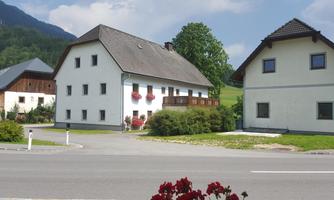 Two residential houses in a rural setting, surrounded by green landscape. In the foreground, red flowers are blooming.