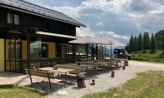 A cozy restaurant in the mountains with several tables and umbrellas. The surroundings are green and hilly, with a partially cloudy sky.