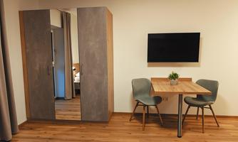 A modern room with a table and two chairs. A television hangs on the wall, and in the background a wardrobe is visible.