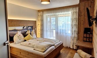 A cozy bedroom with a large wooden bed and floral pillows. Large windows let in plenty of light and offer a view outside.