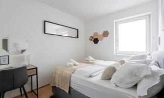 A bright bedroom with a large window and a modern bed. The room is simply decorated and includes a small desk.