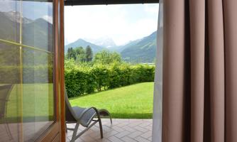 A view from a window of a green meadow and mountains in the background. A deck chair is on the terrace, surrounded by natural beauty.