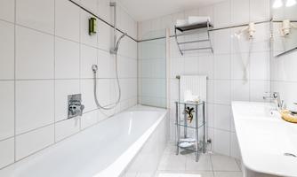 A modern bathroom with a white bathtub and a shower. The walls are tiled and there is a sink with a glass shelf.