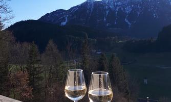 Two glasses of white wine are on a wooden table. In the background, there are majestic mountains and a clear blue sky.
